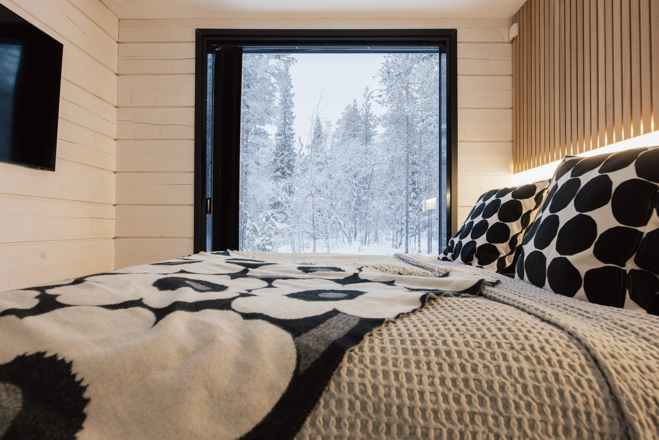A cozy bedroom with a large window showing a snowy forest outside. The bed has a black and white patterned blanket and matching pillows, and the walls are paneled in light wood.