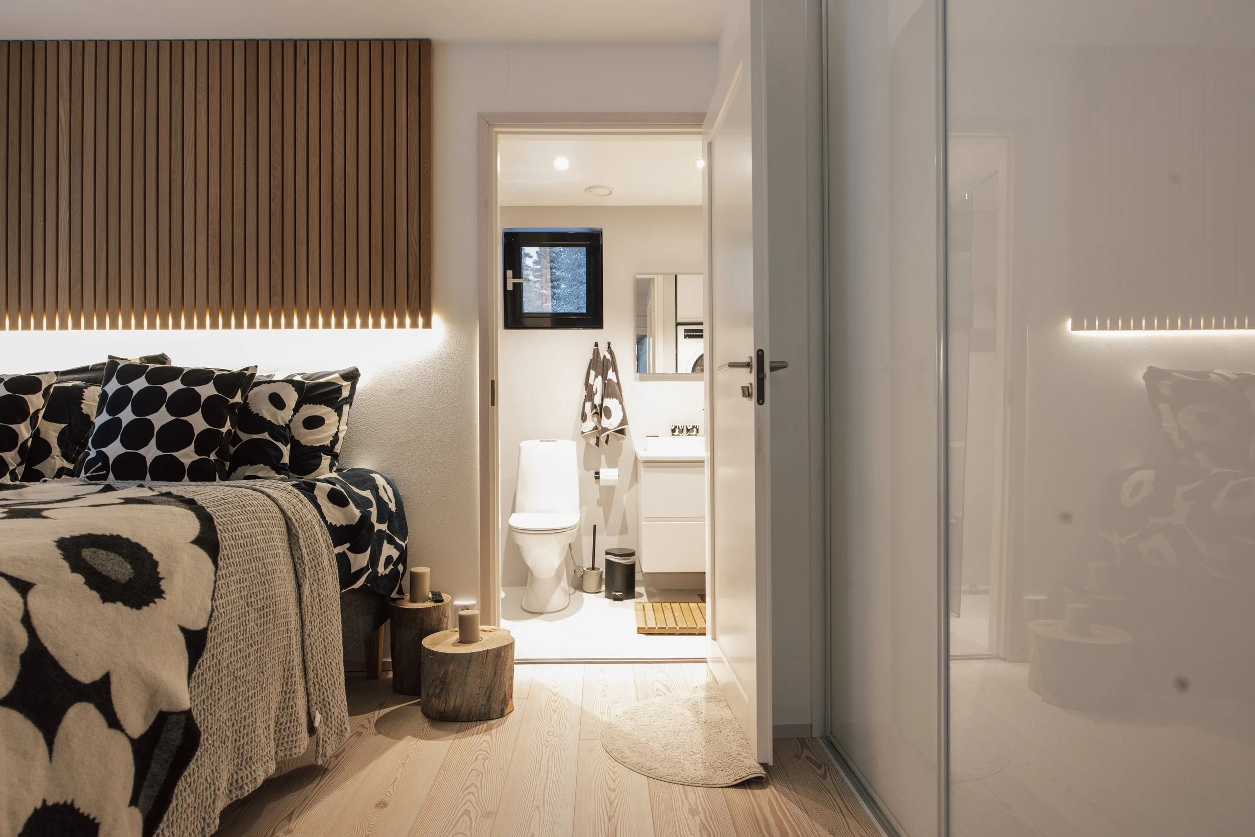 View of a bedroom with a bed on the left with black and white floral bedding, a small wooden side table, and candles. The door to a bathroom is open, showing a white toilet and a small vanity with a mirror. A window and towels are visible in the bathroom. The room has light-colored wooden flooring and a large mirrored closet on the right.