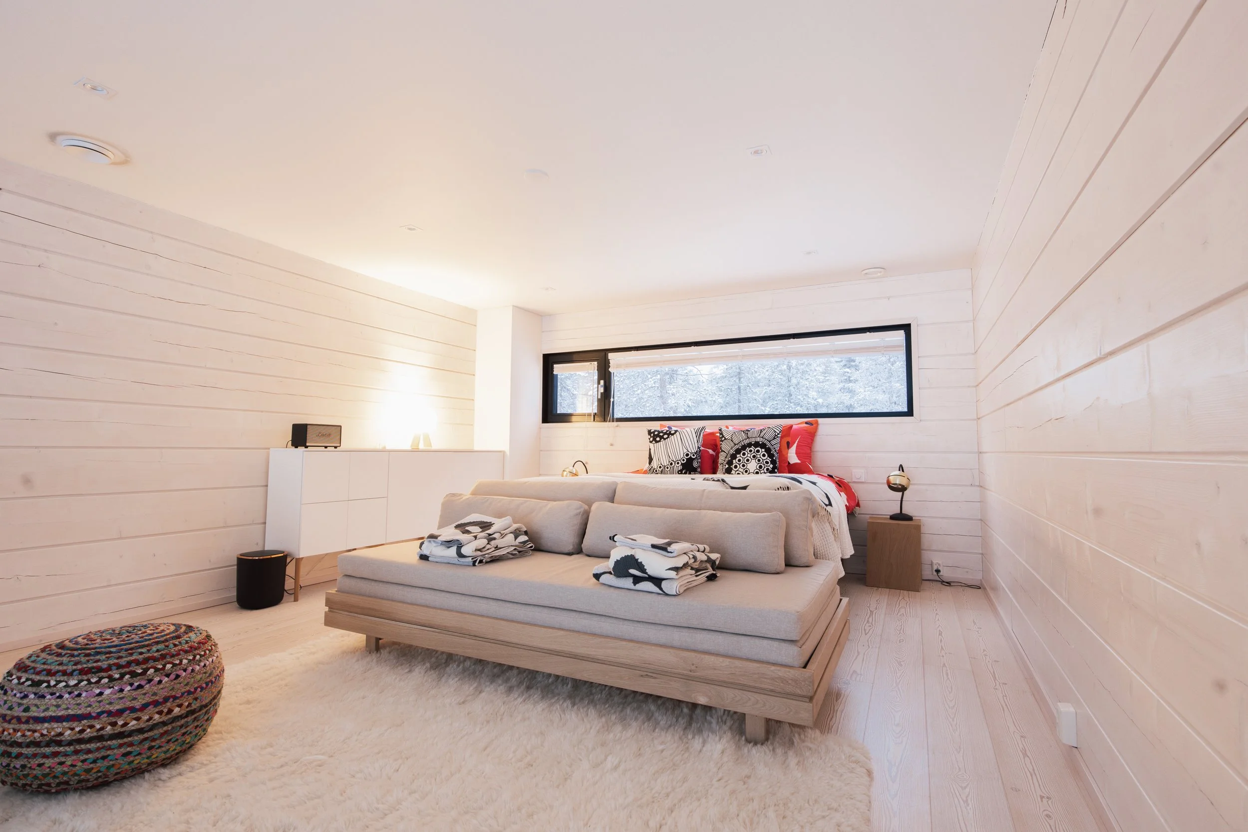 Bedroom with light wood paneled walls, a cream sofa with folded blankets, a bed with patterned pillows, a black horizontal window, a small side table with a lamp, a textured pouf, and a soft rug.