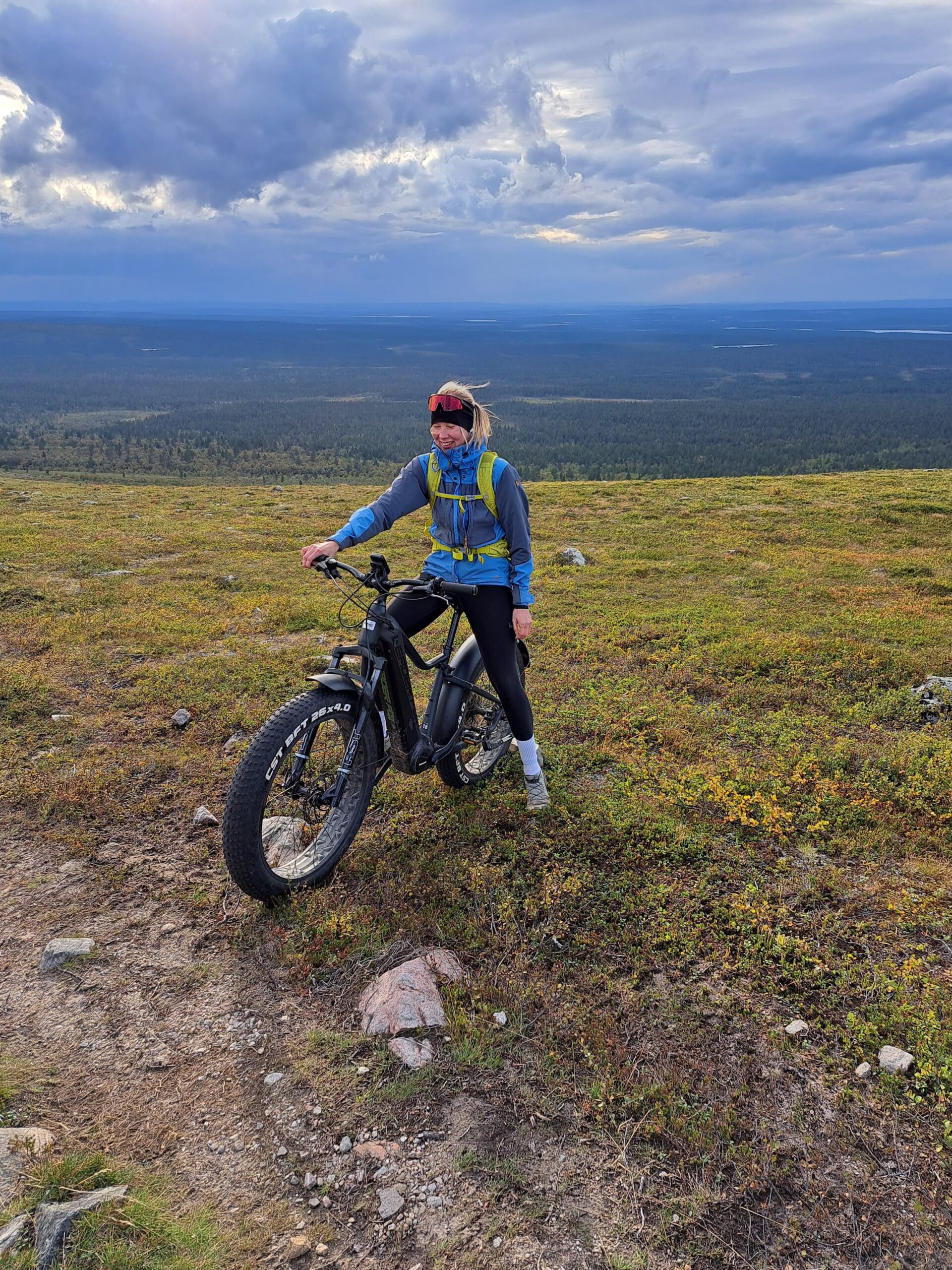 A woman stands with a mountain bike on a grassy hilltop overlooking a vast forest and cloudy sky.