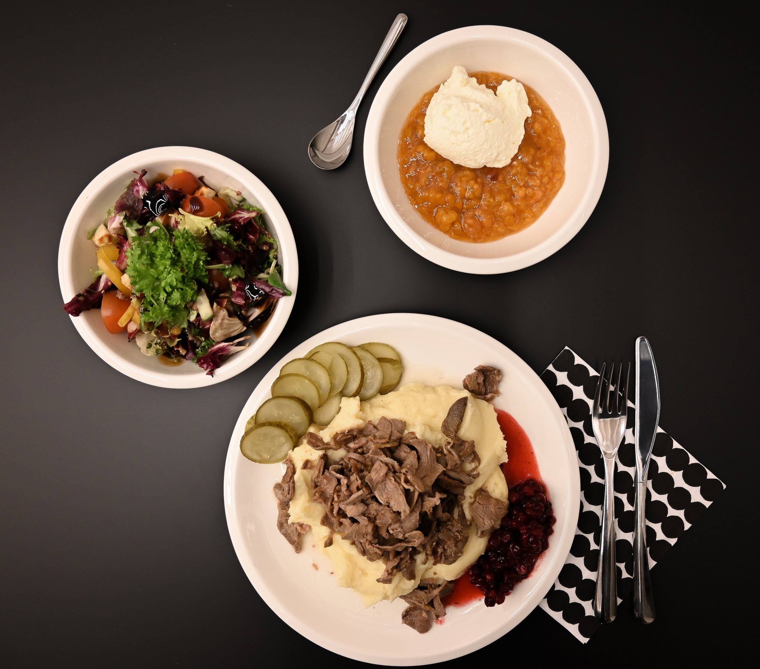 A plate with mashed potatoes topped with sliced pickles, shredded beef, cranberry sauce, and a dollop of red sauce. A bowl of mixed vegetable salad with greens, cherry tomatoes, and herbs. A bowl of pea soup with bread on top. Silverware and a black and white polka dot napkin rest beside the plate.