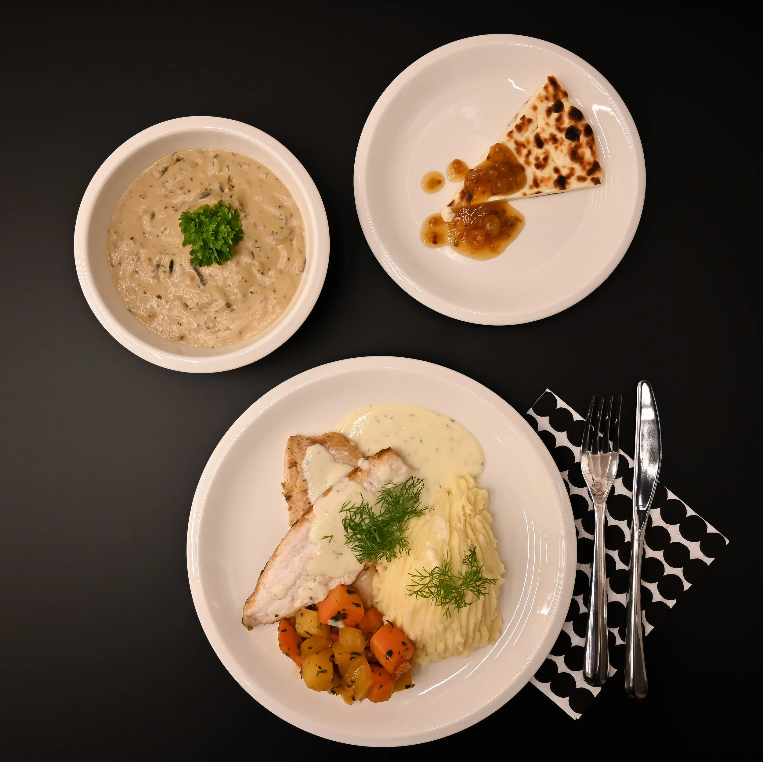 Plate with mashed potatoes, grilled chicken breast with sauce, and seasoned vegetables; bowl of creamy soup garnished with parsley; small plate with a slice of flatbread and curry sauce; black table with silverware on a patterned napkin.