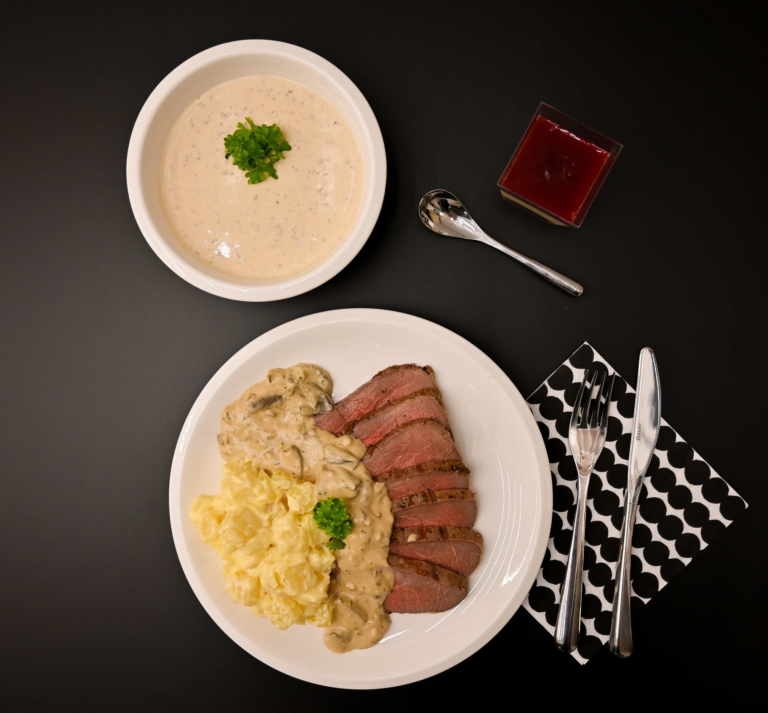 Plate with sliced steak, mashed potatoes, and mushroom gravy, served with creamy mushroom sauce. Soup with parsley garnish, a glass of red wine, and a set of silverware on a black and white napkin. Black table surface.