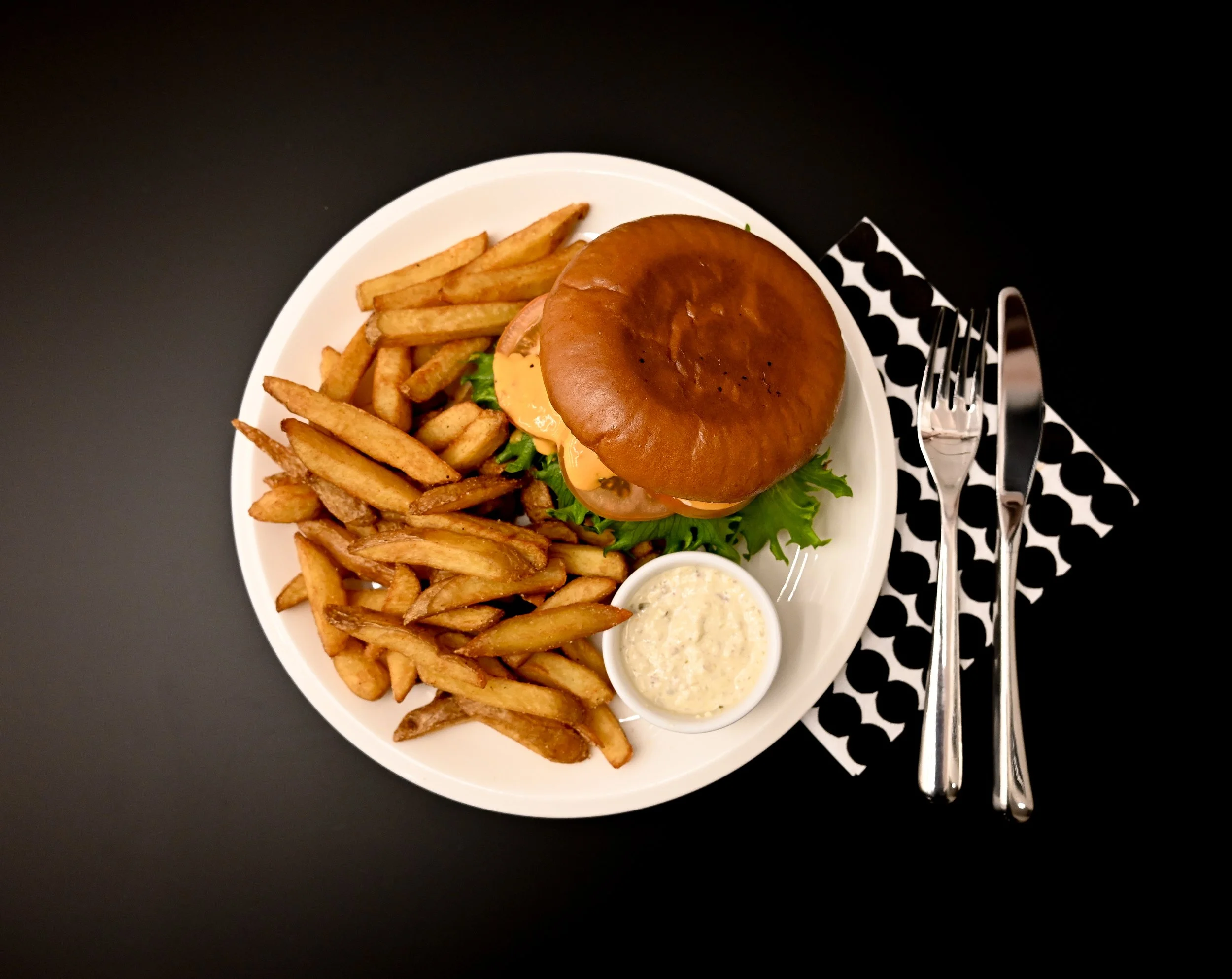 Plate with cheeseburger, French fries, and a small bowl of sauce on a black table with a black and white napkin and silverware.