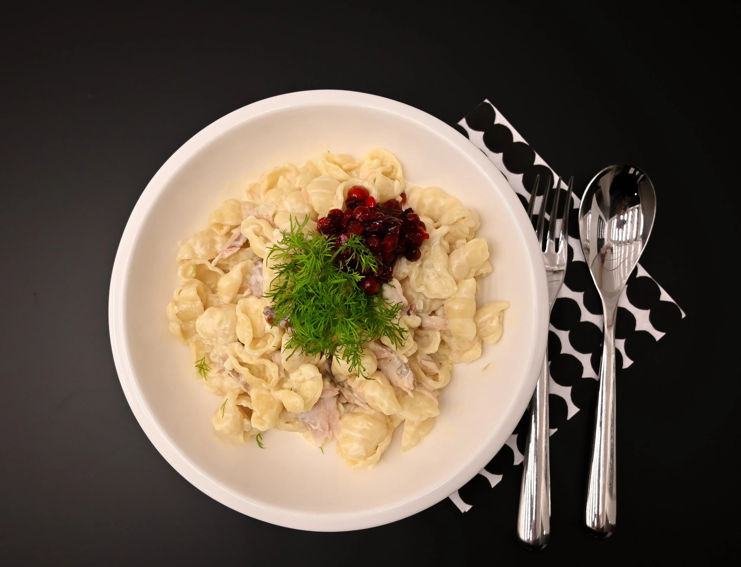 A bowl of creamy pasta garnished with herbs and berries, served with cutlery on a black and white napkin on a black surface.