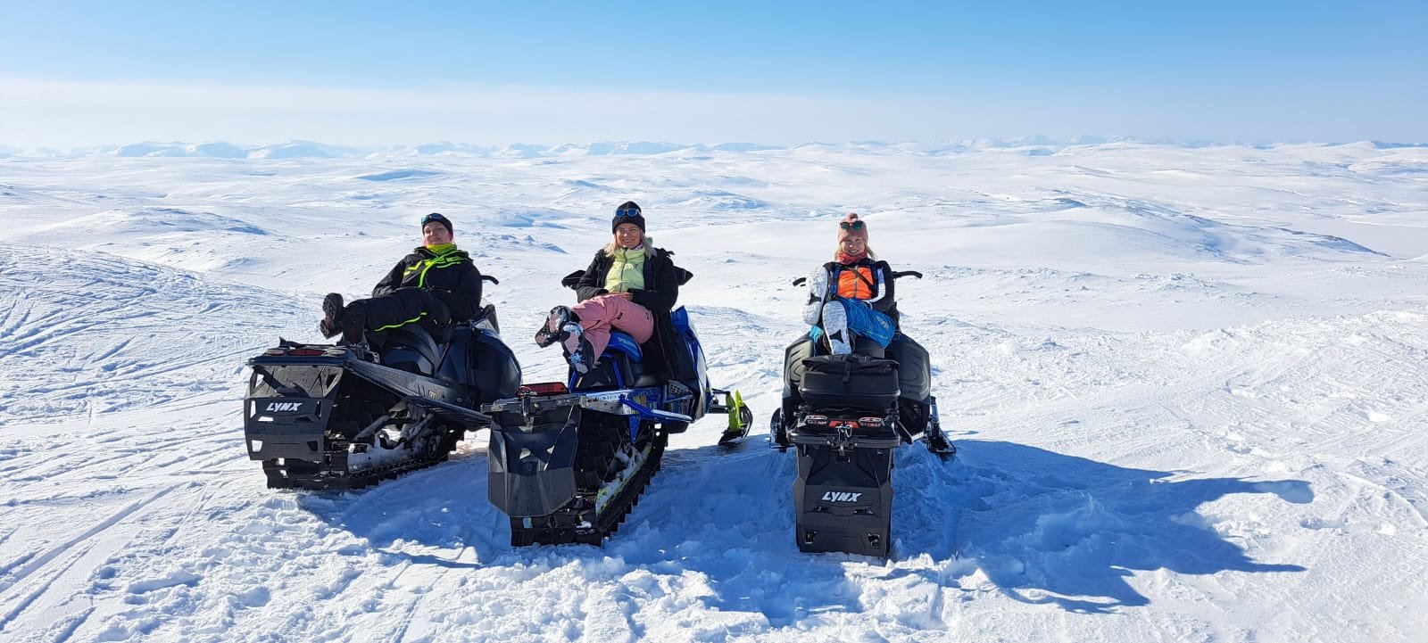 Three people sitting on snowmobiles in a snowy landscape with clear blue sky.