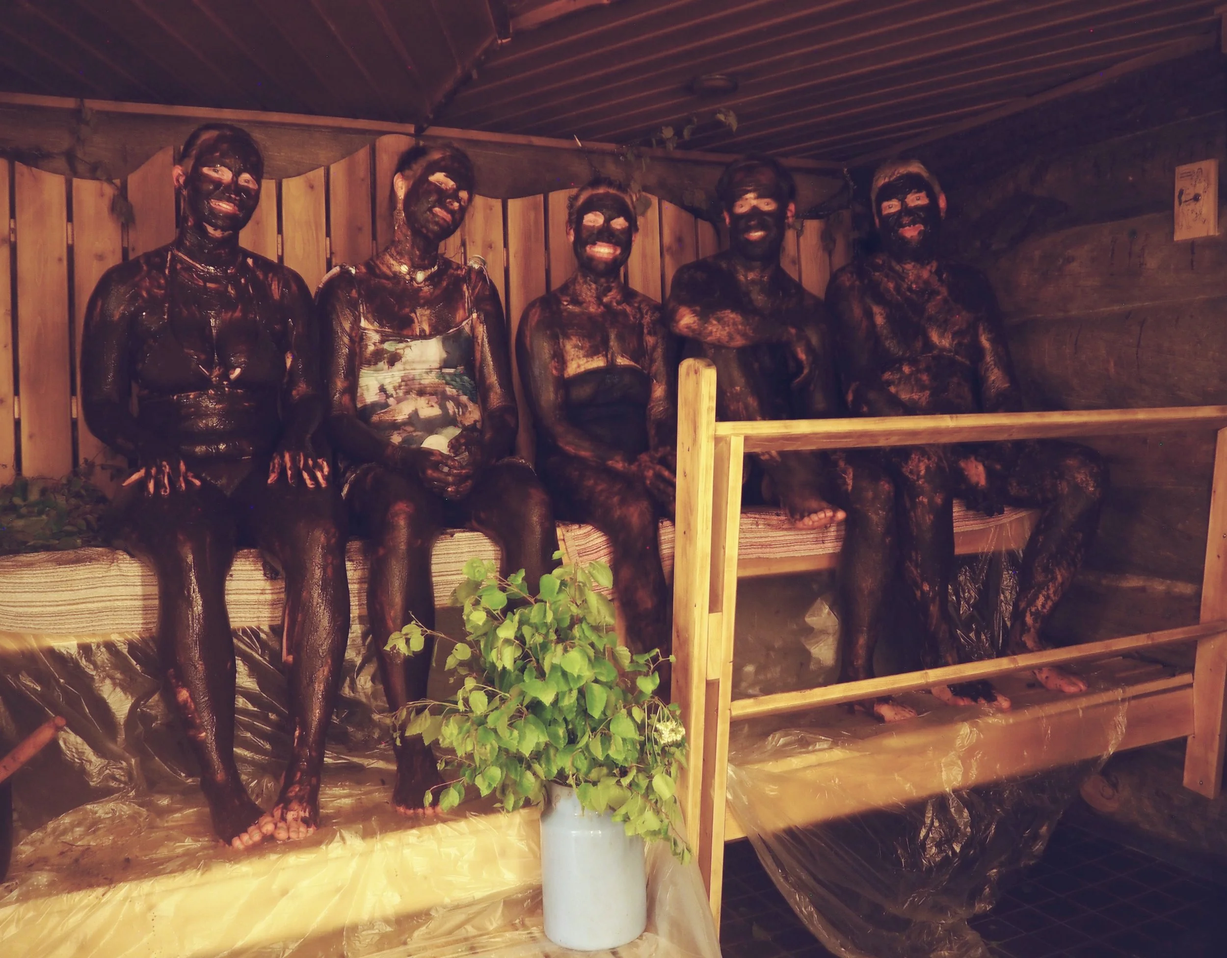 Group of five people covered in dark mud, sitting on a wooden bench inside a sauna or cabin, smiling. There is a plant in a white pot in the foreground.