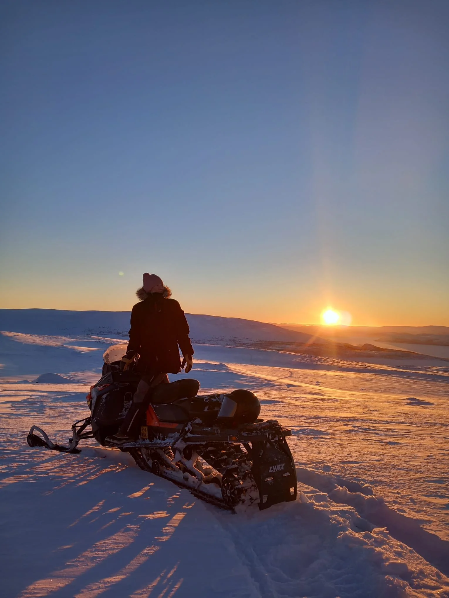 A person dressed in winter clothing standing beside a snowmobile on a snow-covered landscape during a sunset or sunrise.