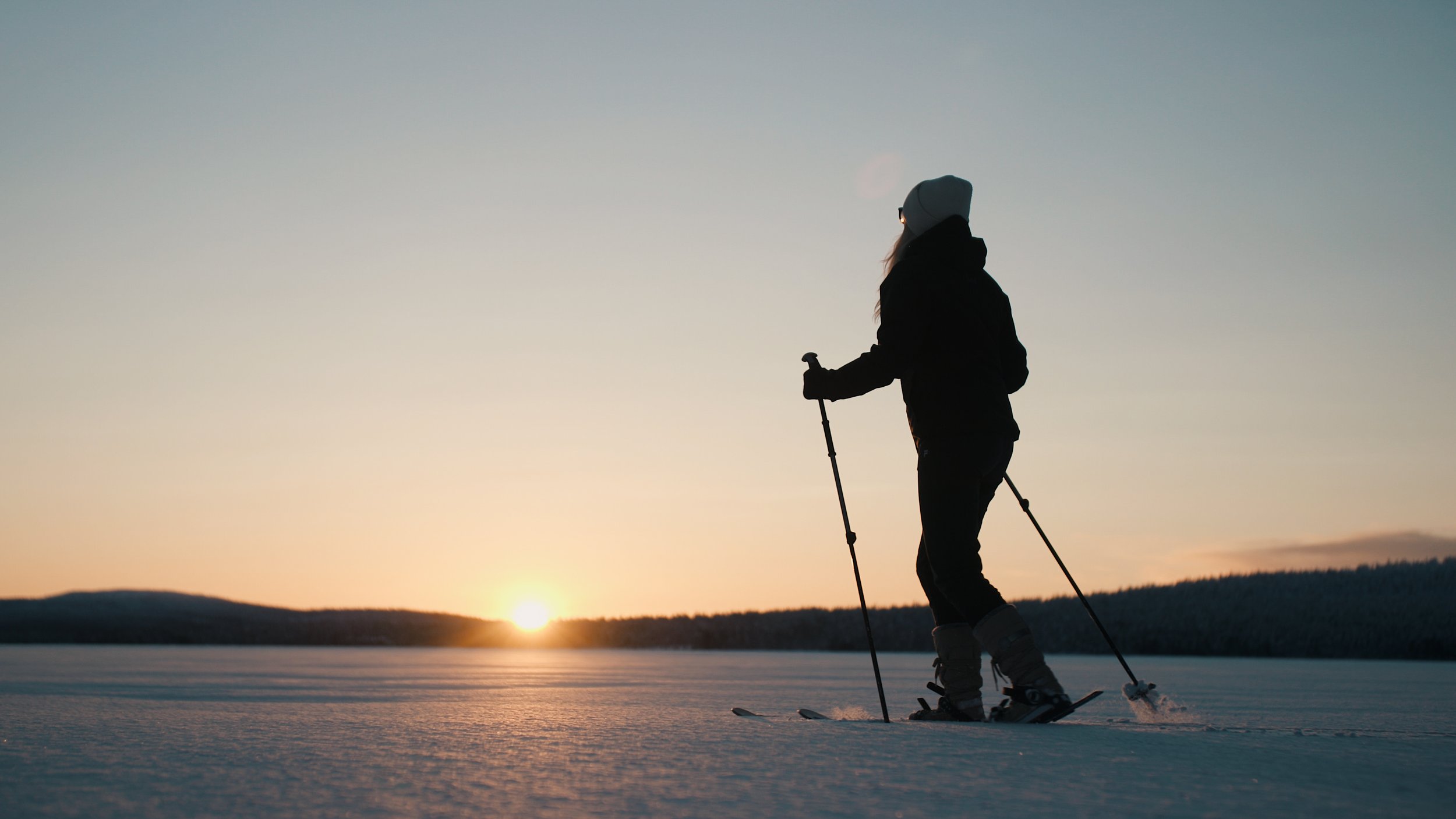 Silhouette of a person cross-country skiing on a snow-covered landscape during sunset.