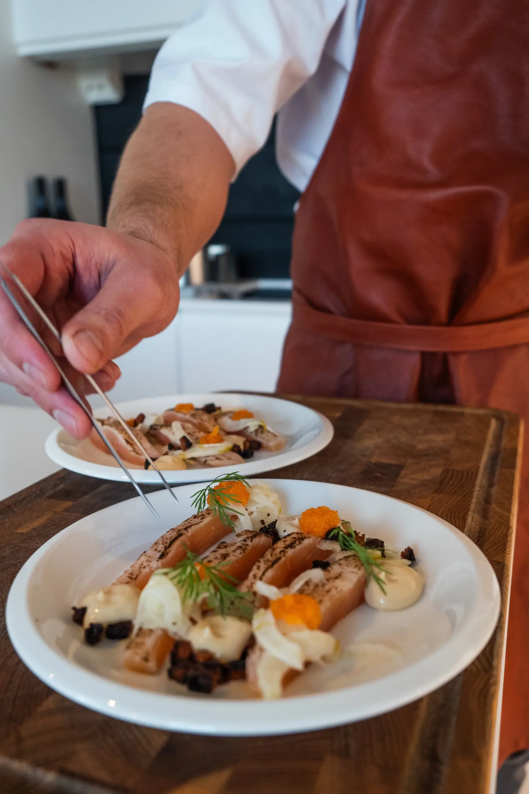 A chef garnishing plates of gourmet dish with tweezers, featuring sliced fish, herbs, and garnishes on white plates.