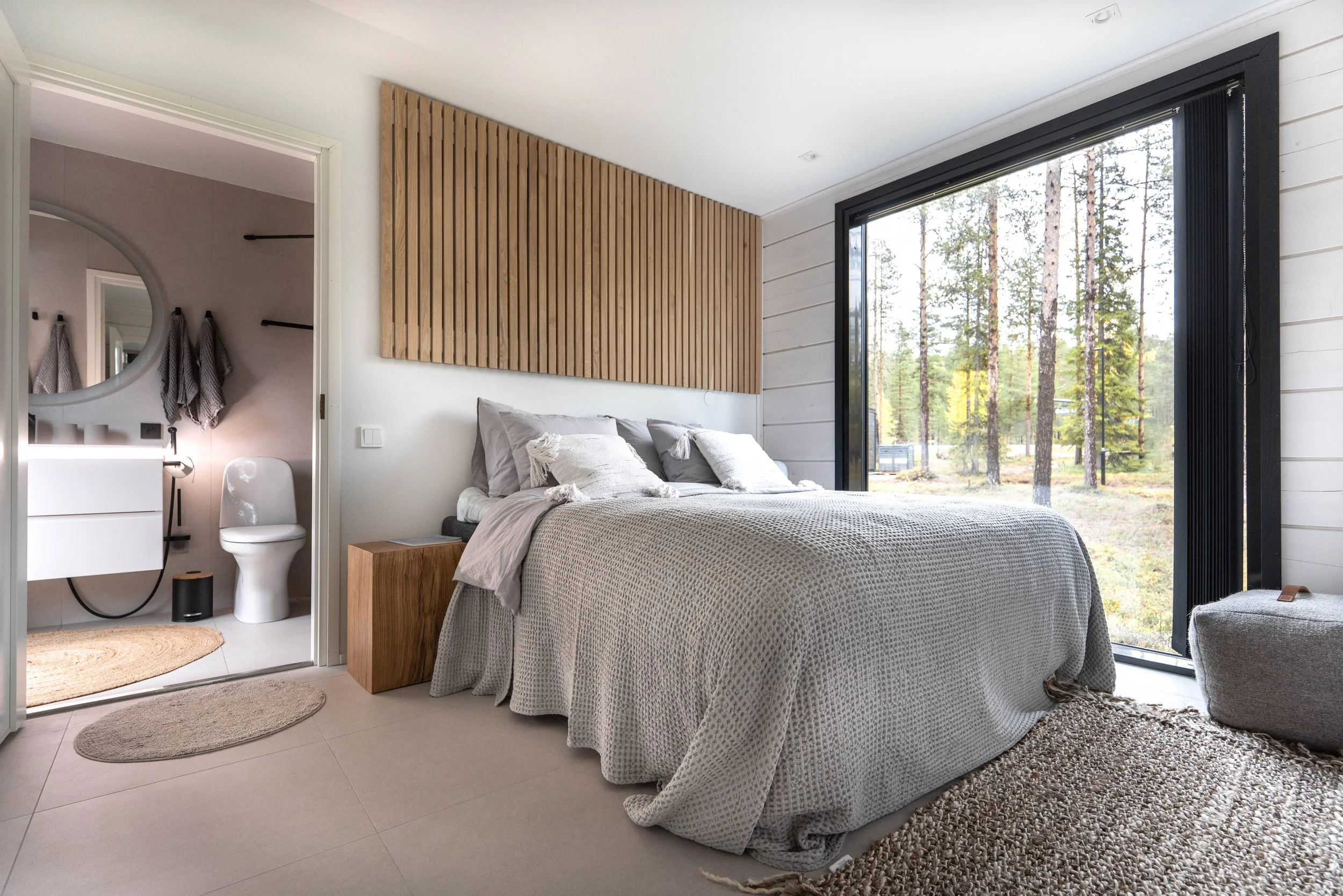 A modern bedroom with a large bed covered in a light gray textured blanket, six pillows, and a wooden headboard on the wall. Large sliding glass doors reveal a wooded outdoor scene. An open door shows a bathroom with a round mirror, a white sink, and a toilet.