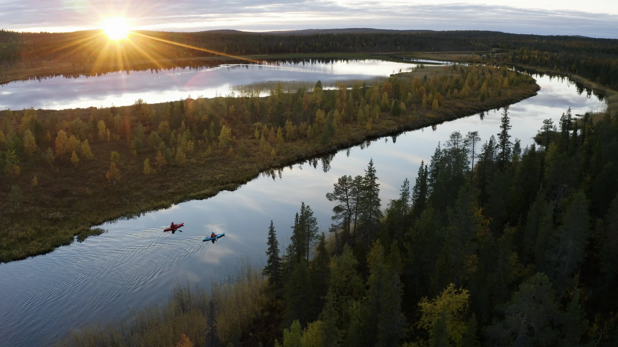 Two kayakers paddle through a calm river surrounded by a lush forest with trees changing colors, under a cloudy sky with the sun setting on the horizon.