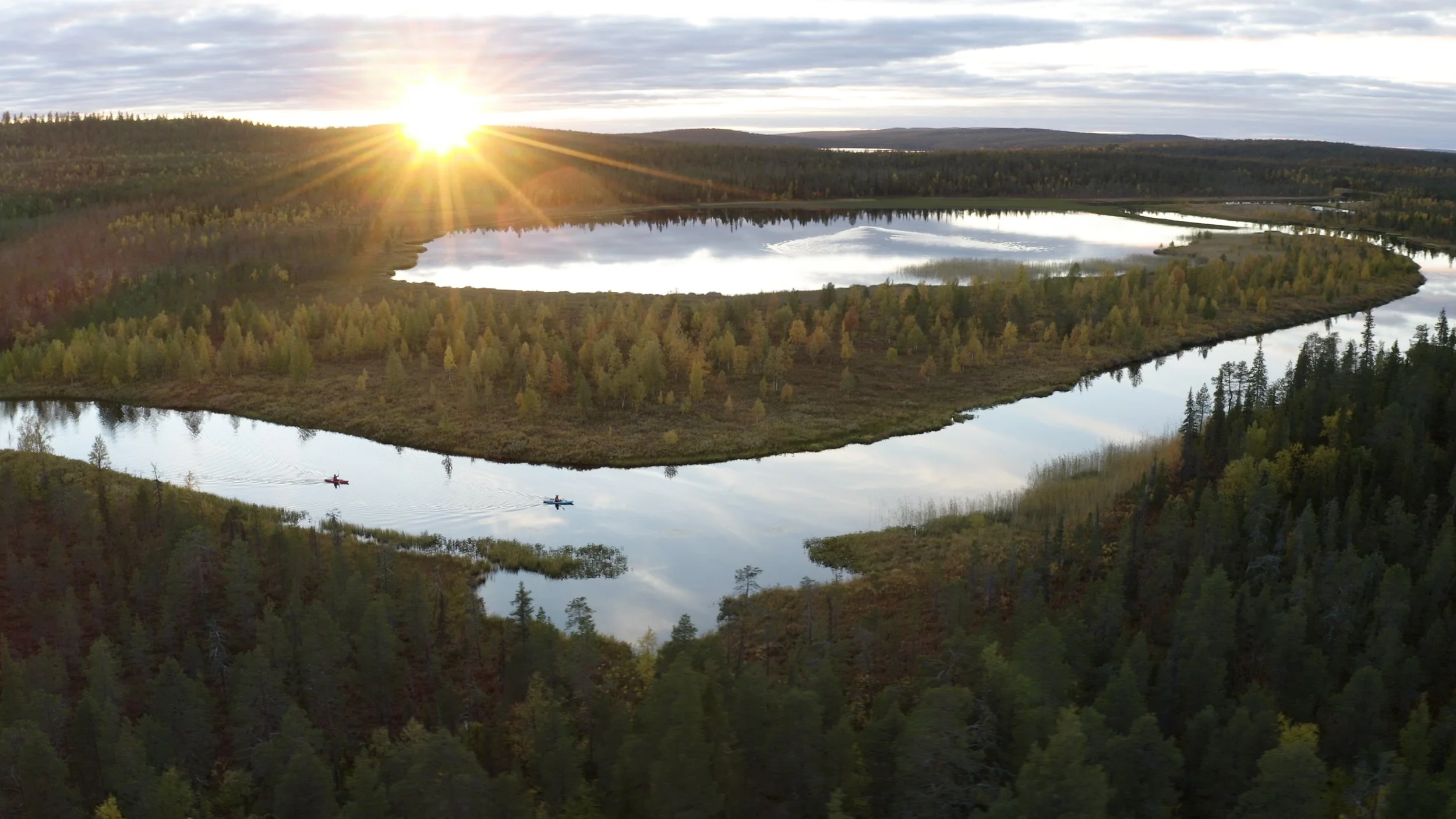 Two kayakers paddling on a calm lake reflecting the sky and surrounding trees, with a sunset over a forested landscape in the background.