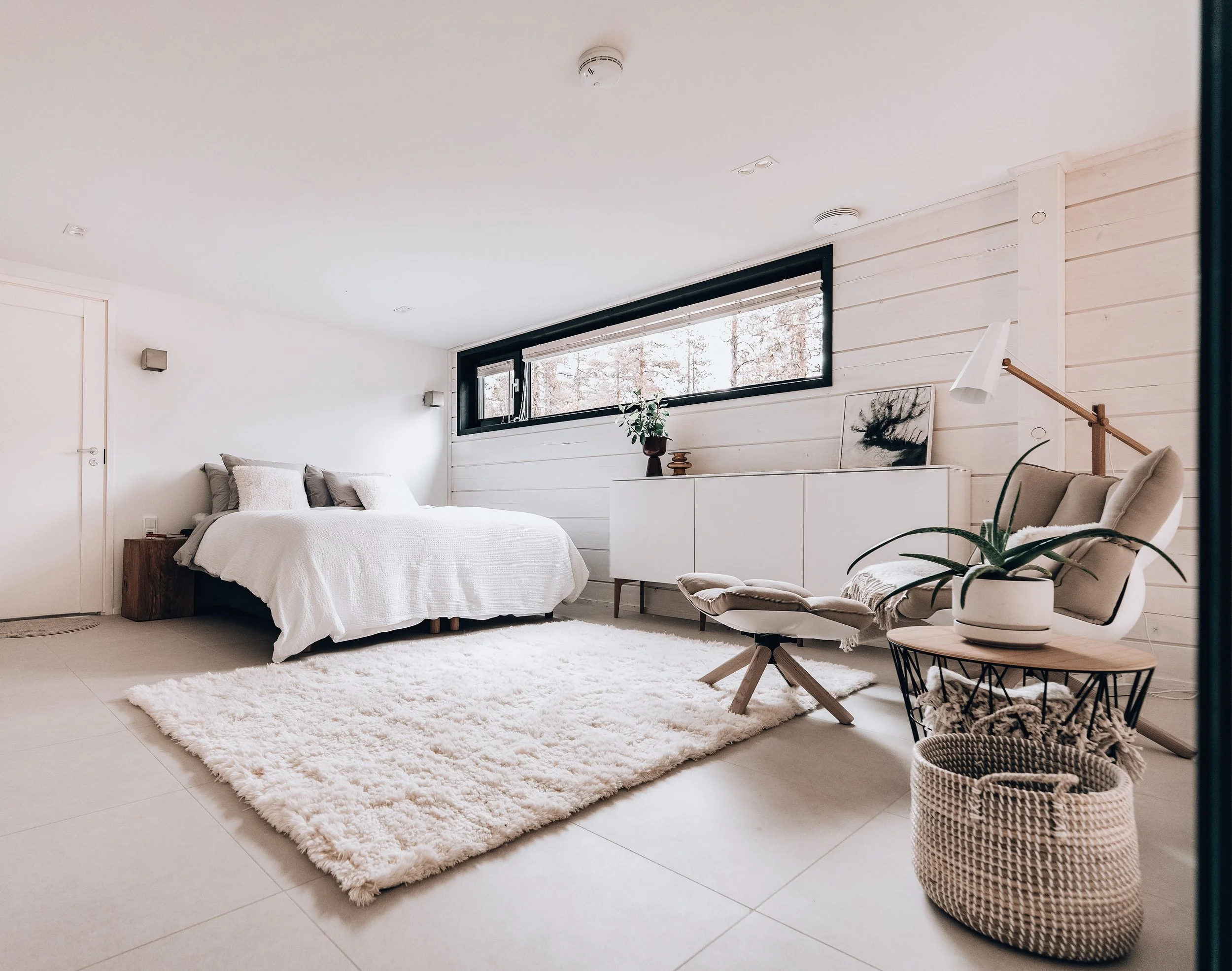 A bright, minimalist bedroom with white walls, a large bed with white bedding, a wooden bedside table, a plush white rug, a white dresser with decorative items, a beige armchair with an ottoman, a side table with a potted plant, and a black-framed horizontal window showing trees outside.