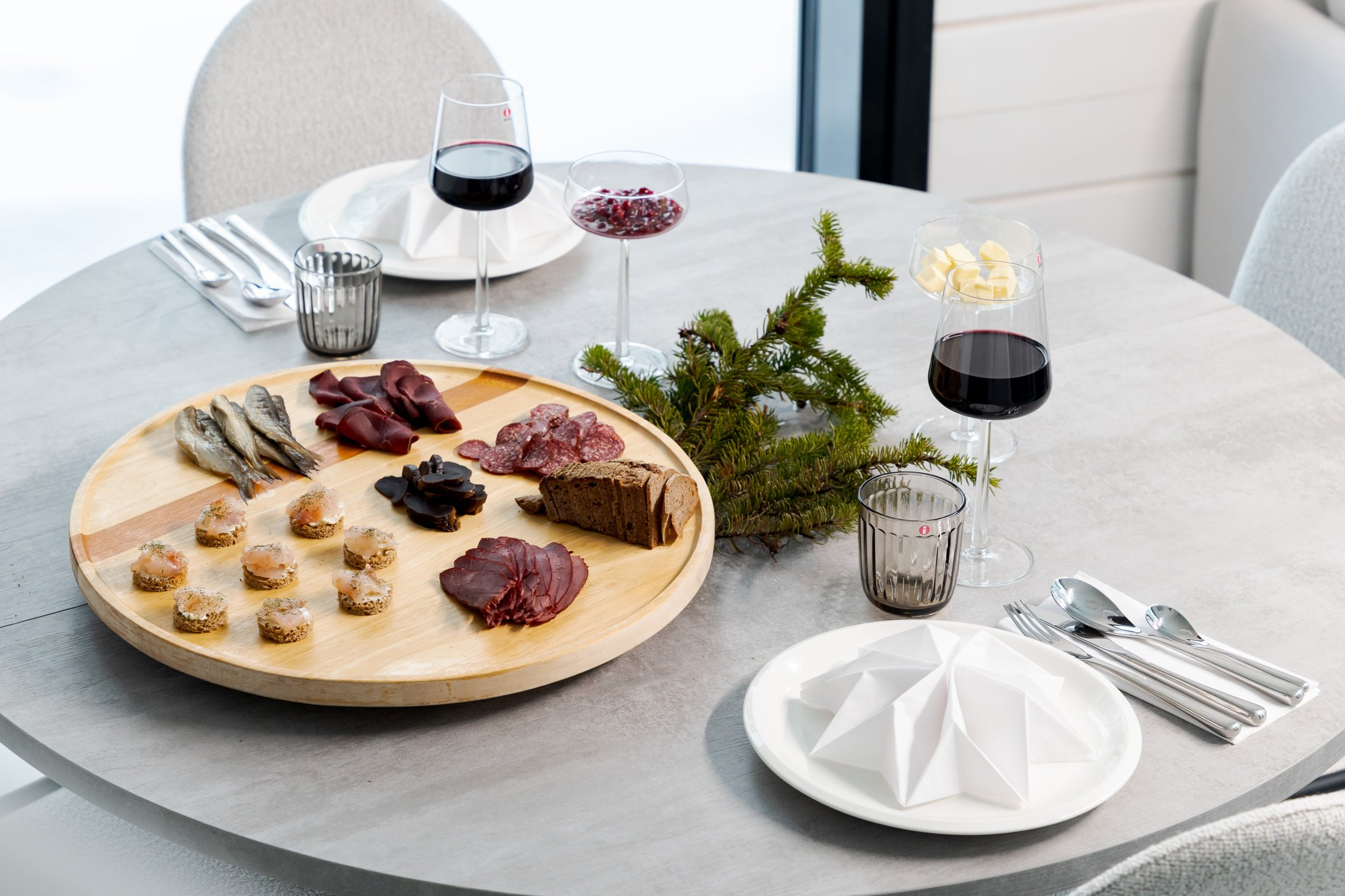 A round table set for a meal with a charcuterie board, glasses of red wine, and decorative greenery.