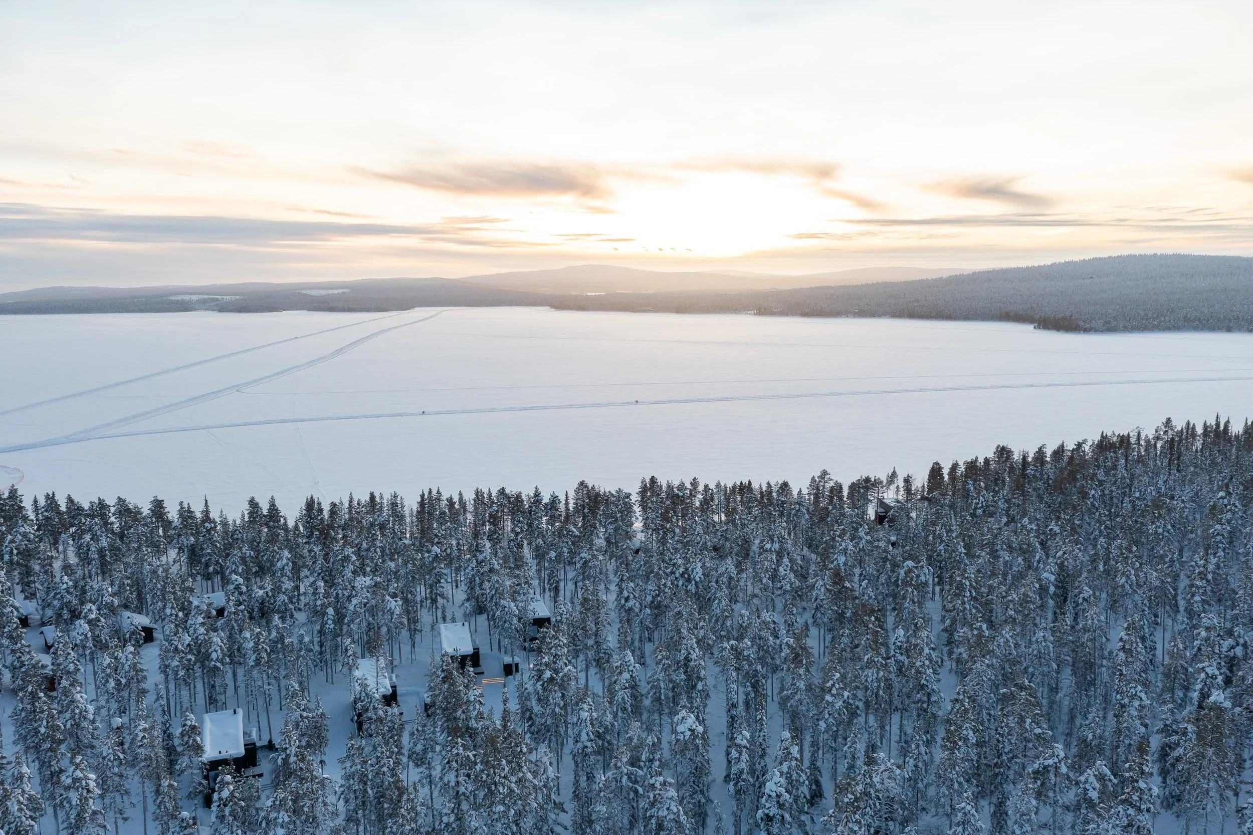 A snowy landscape with a pine forest in the foreground and a frozen lake in the background, with the sun setting or rising in the sky.