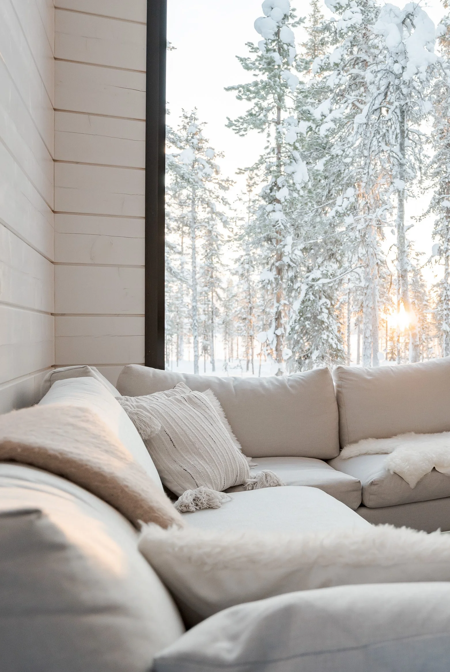 Cozy white sectional sofa with pillows and throws inside a cabin with large window showing snow-covered trees and sunlight outside.