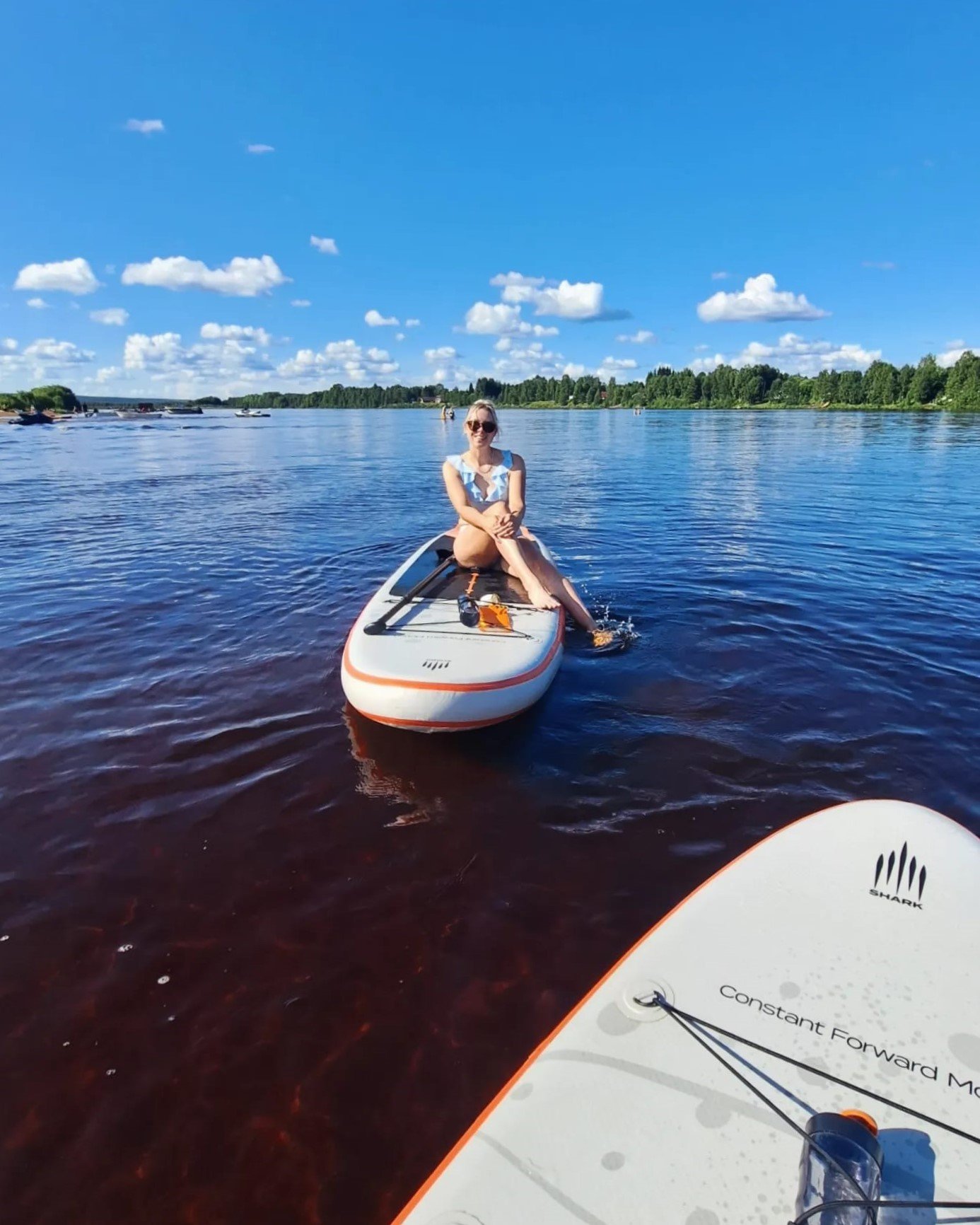 A woman sitting on a paddleboard in a body of water, smiling and wearing sunglasses, with a clear blue sky and green trees in the background.