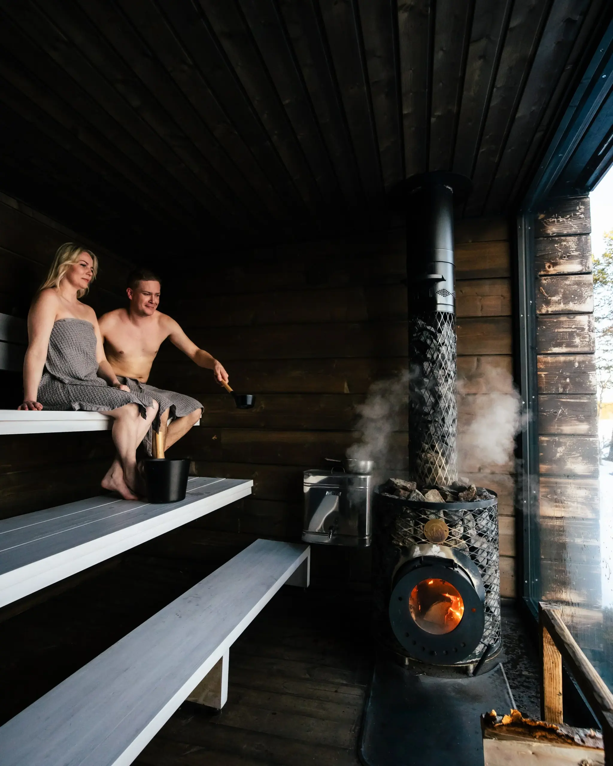 A man and woman in towels sitting on wooden benches inside a sauna, with the man pouring water on a stove for steam.