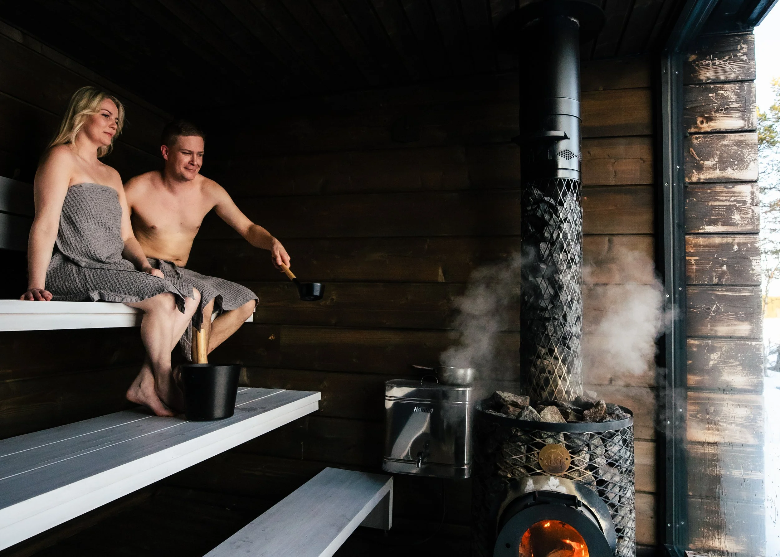 Two people sitting on a wooden sauna bench, wrapped in towels, with one pouring water onto a hot wood-burning sauna stove emitting steam in a sauna room.