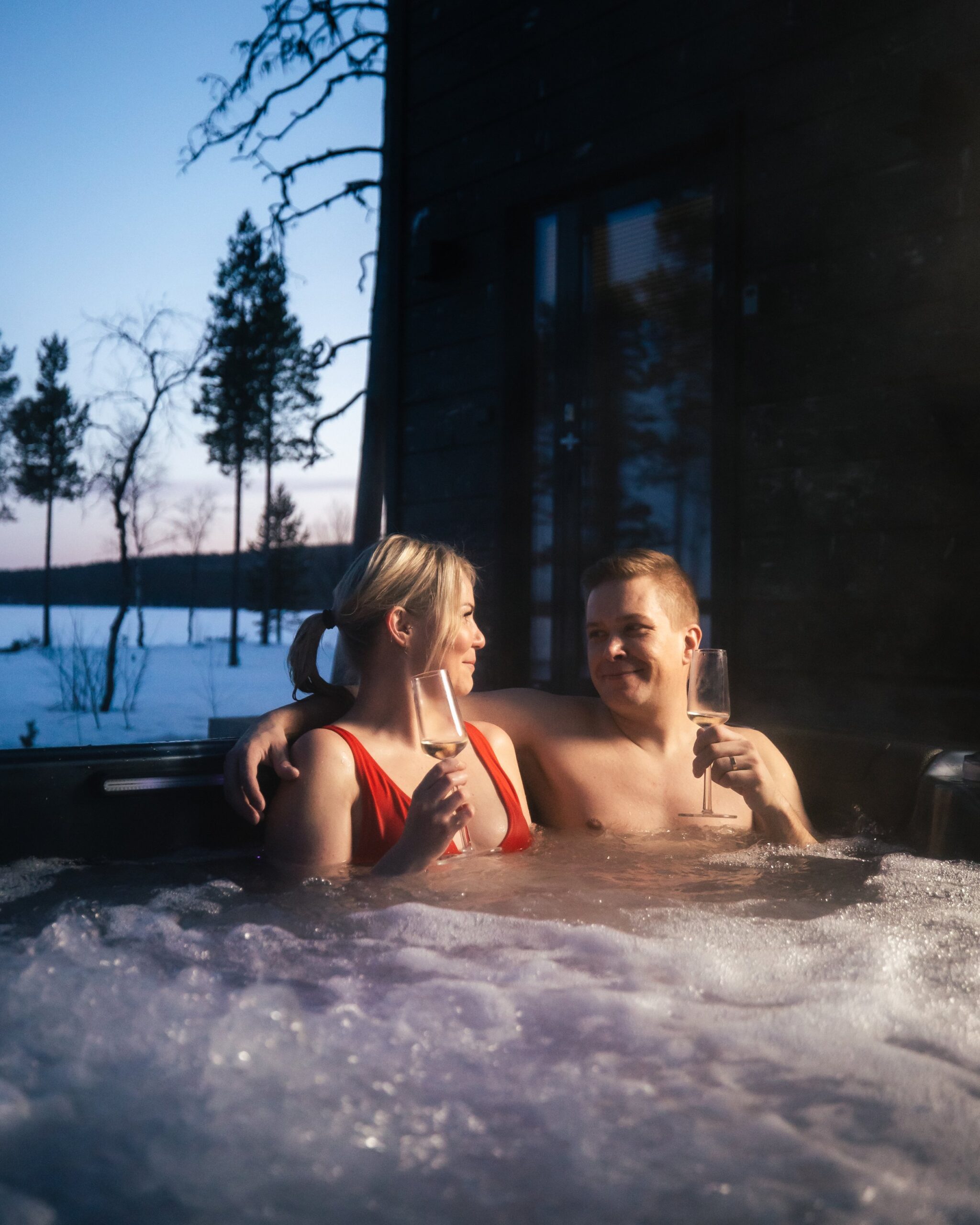 A couple relaxing in a hot tub outdoors in winter, holding glasses of wine, with snow and trees in the background.