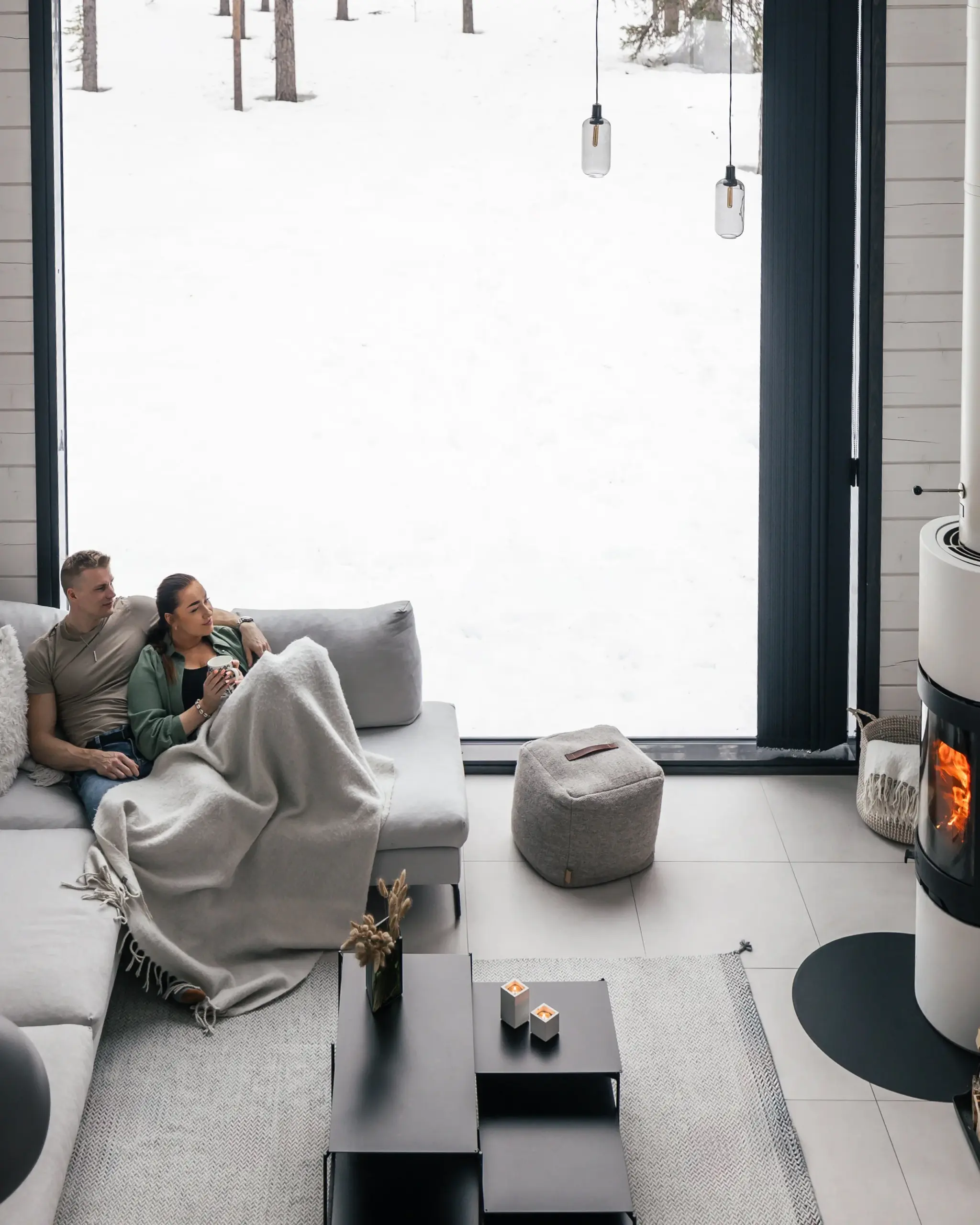 Living room with a large window overlooking a snowy landscape, two people sitting on a sofa covered with a blanket, a black coffee table, a candle holder, a fireplace with a fire, and a pouf.