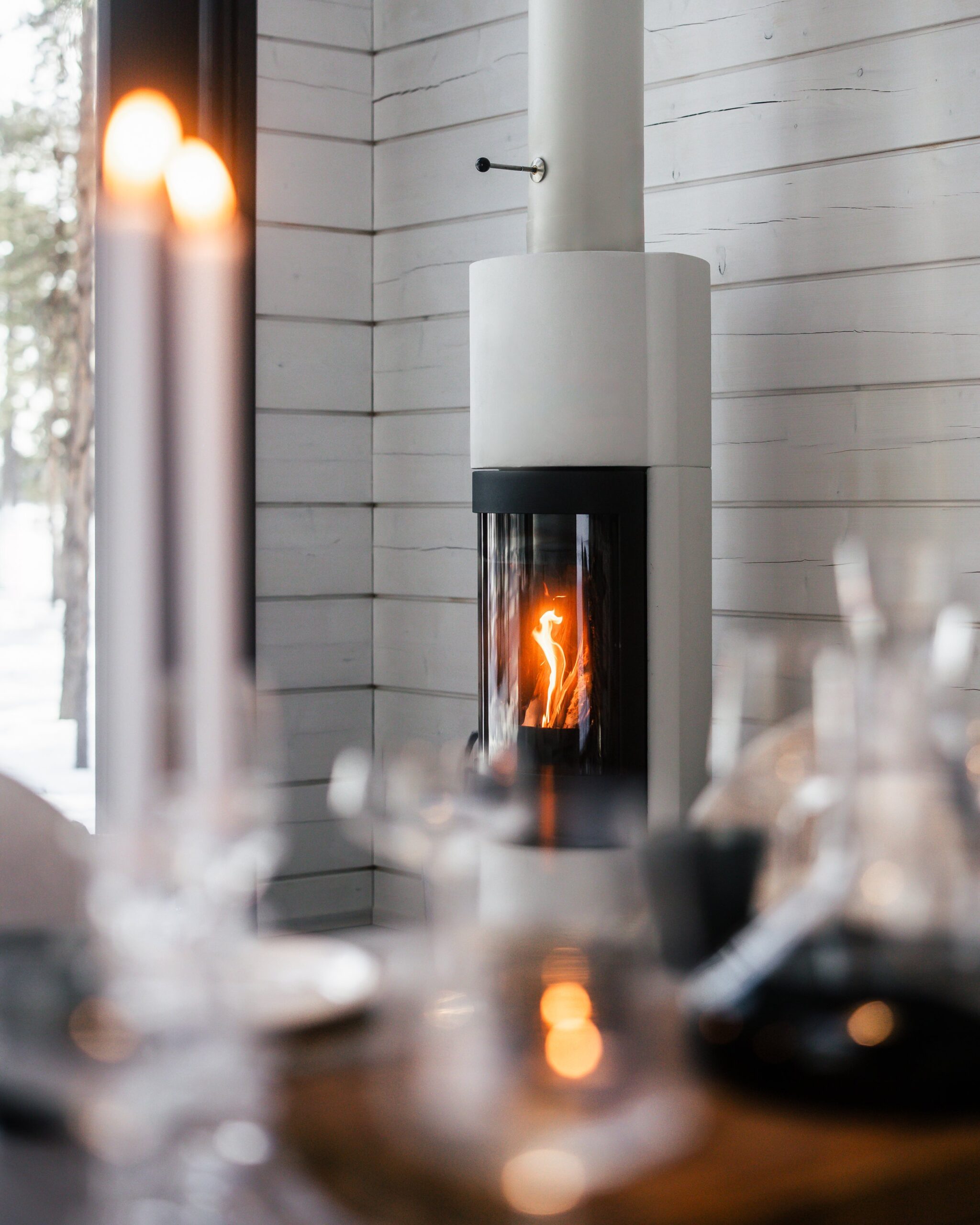 A modern white and black wood stove with a glass window showing a flickering flame, set against a light wood-paneled wall, with blurred candles and tableware in the foreground.