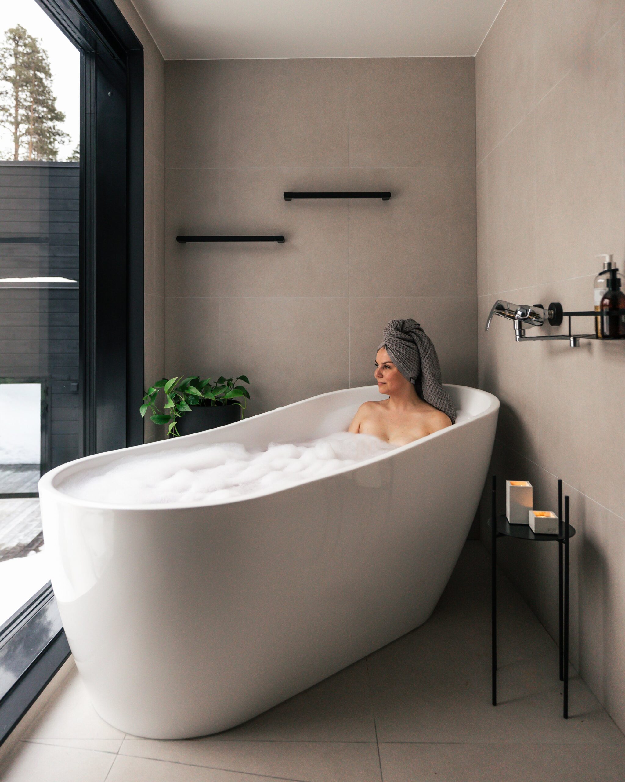 Woman relaxing in a modern bathtub with bubbles, wrapped in a towel on her head, next to a large window with a snowy outdoor scene.