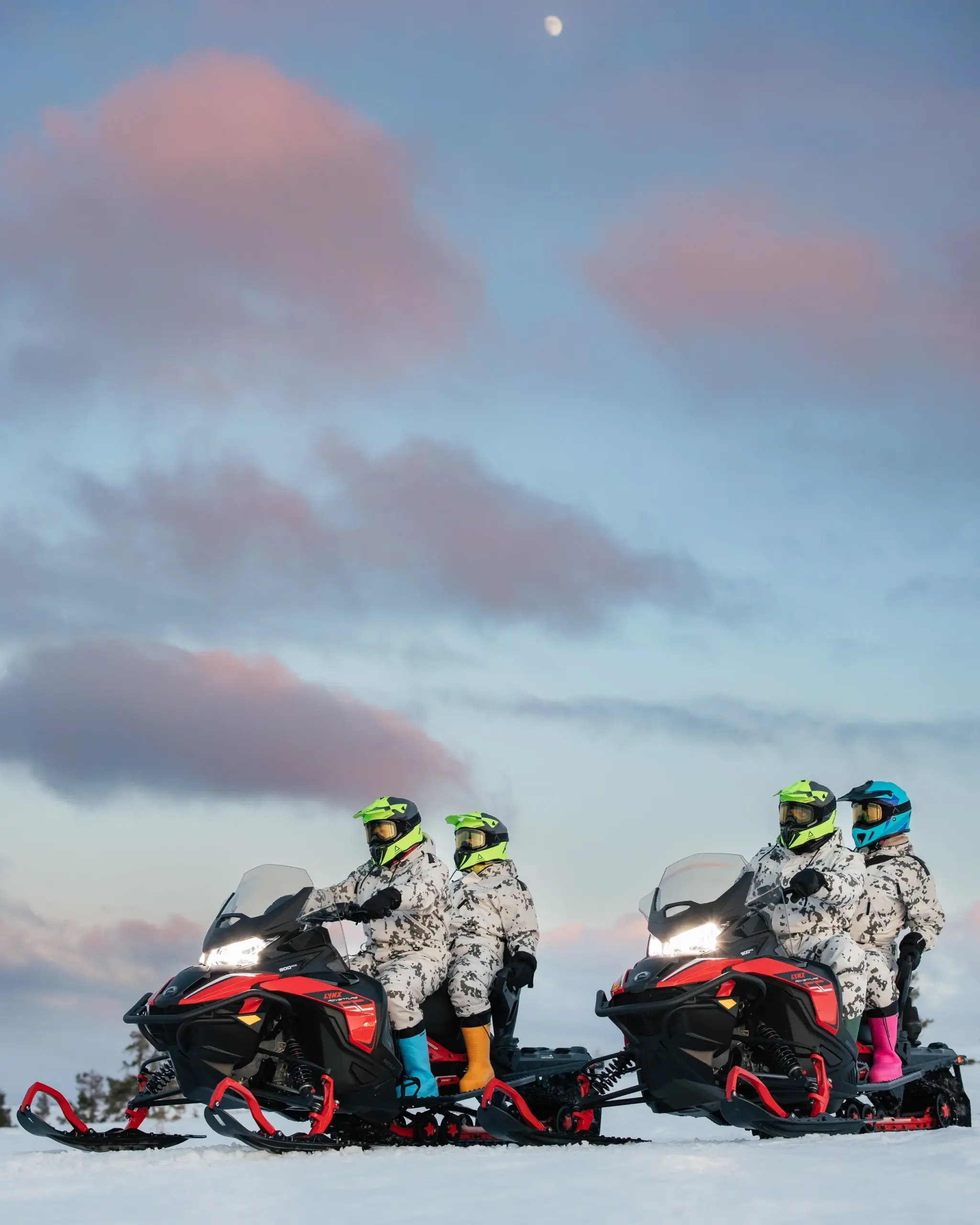 Four people riding snowmobiles on snow, wearing camouflage outfits and helmets with neon accents, under a partly cloudy sky with the moon visible.