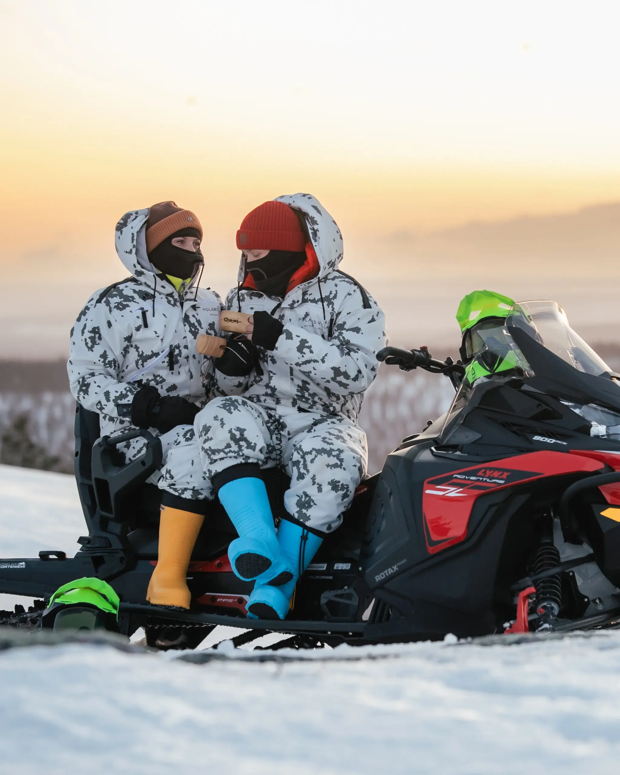 Two people in winter gear sitting on a snowmobile in a snowy landscape at sunset, wearing camouflage suits, hats, and masks, with one person showing something on a device.