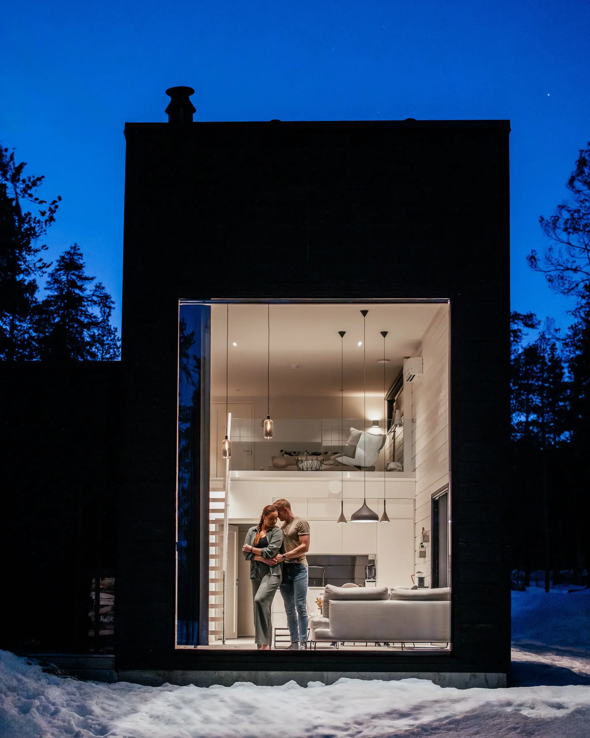 A modern two-story house with large window at night. Inside, a couple is standing together on the first floor, illuminated by warm lighting, with a white sofa and kitchen visible in the background. Snow covers the ground outside.