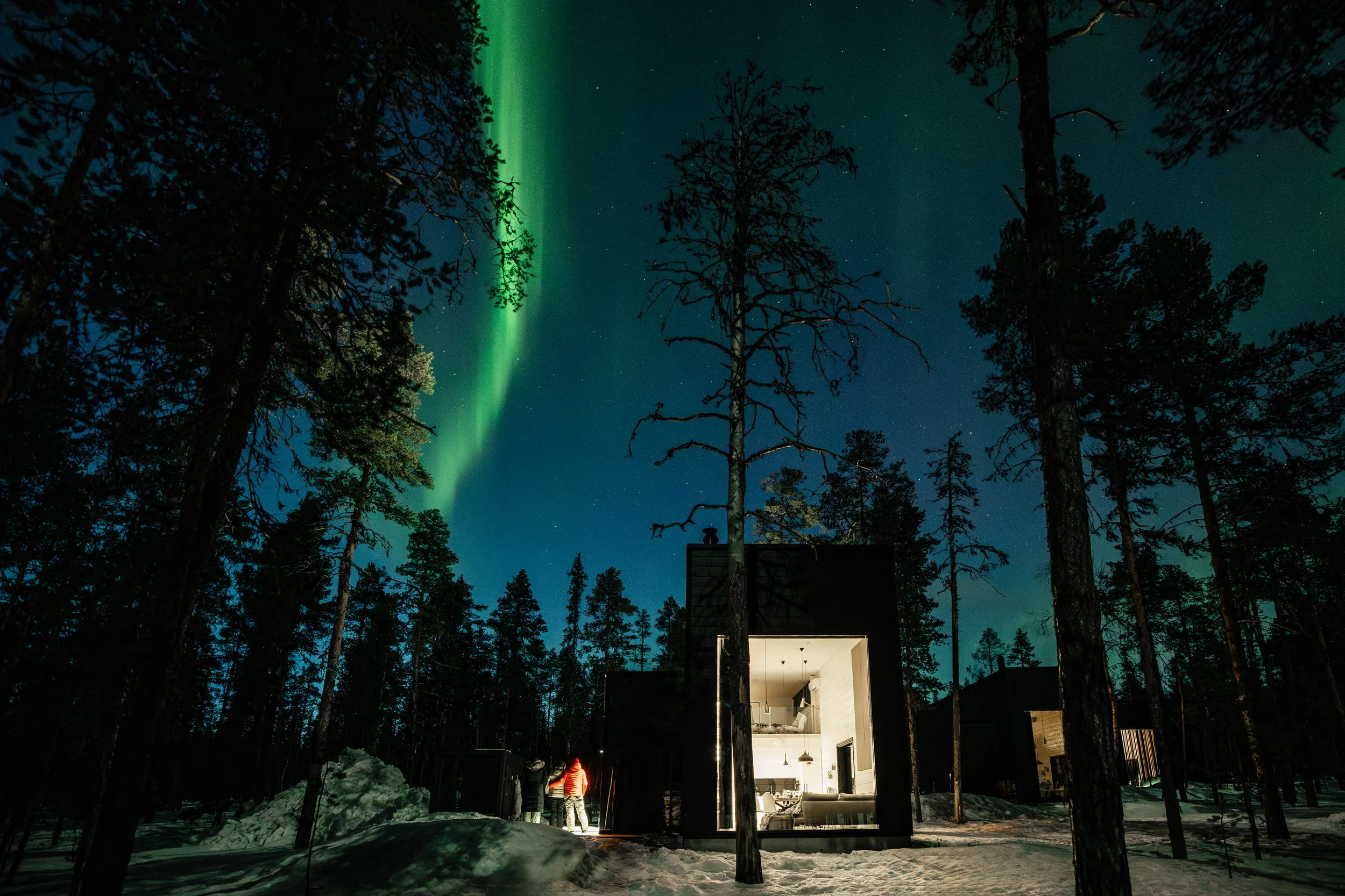 Nighttime in a snowy forest with a house lit from inside. The northern lights and stars are visible in the sky, with a green aurora stretching upward.