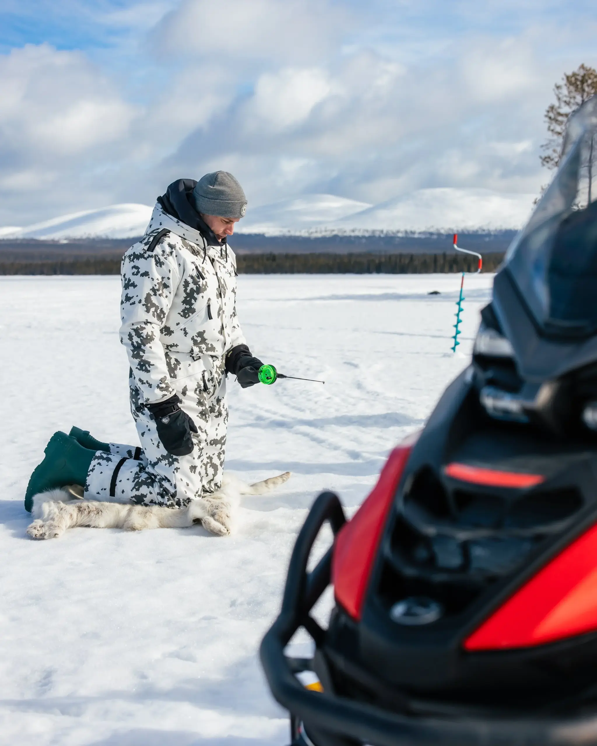 A person kneeling on snow in winter clothing, holding a specialized tool in snowy landscape with mountains in the background, a dog lying on the snow nearby, and a snowmobile in the foreground.