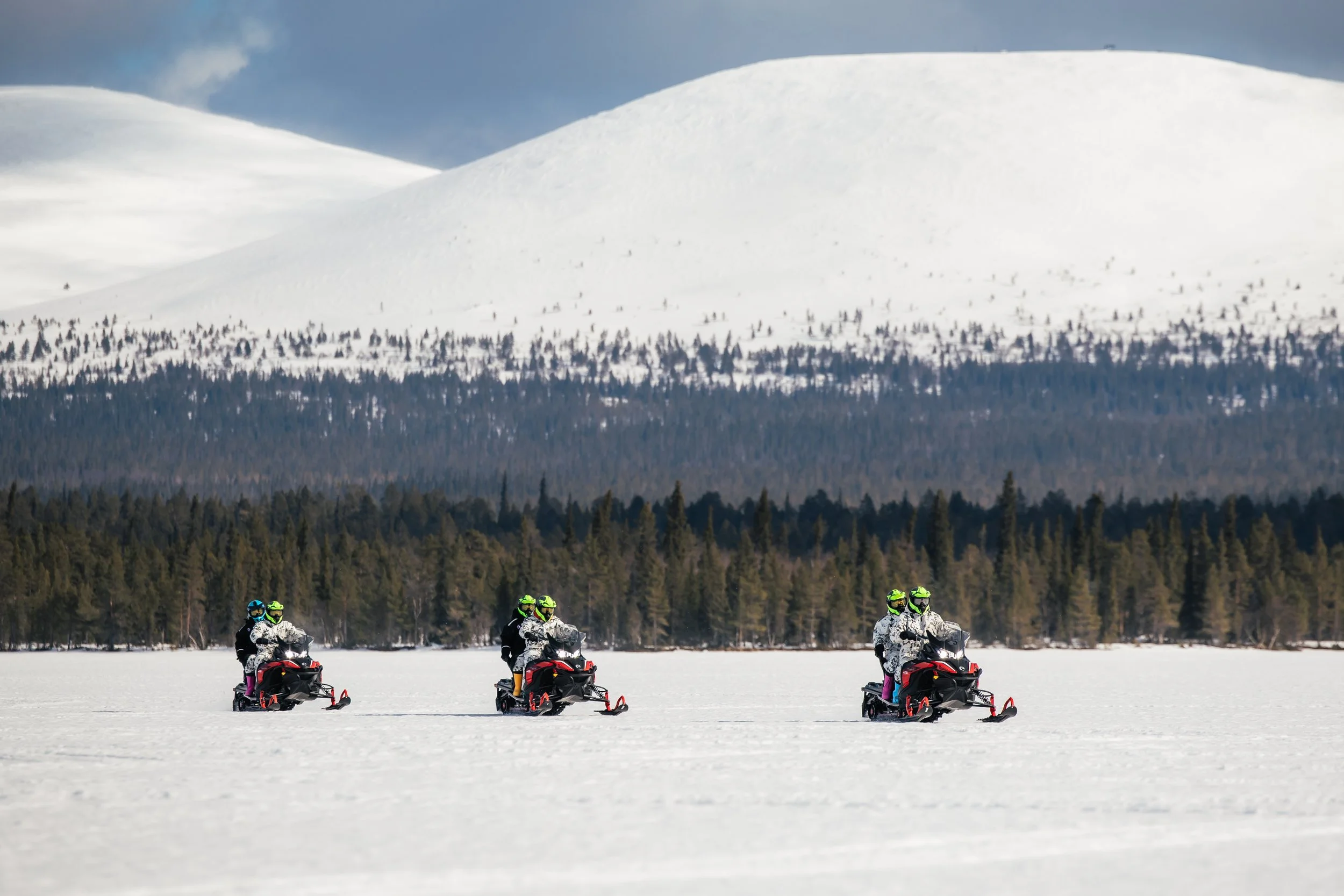 Three people riding snowmobiles on a snow-covered field with a forest and snow-capped mountains in the background.